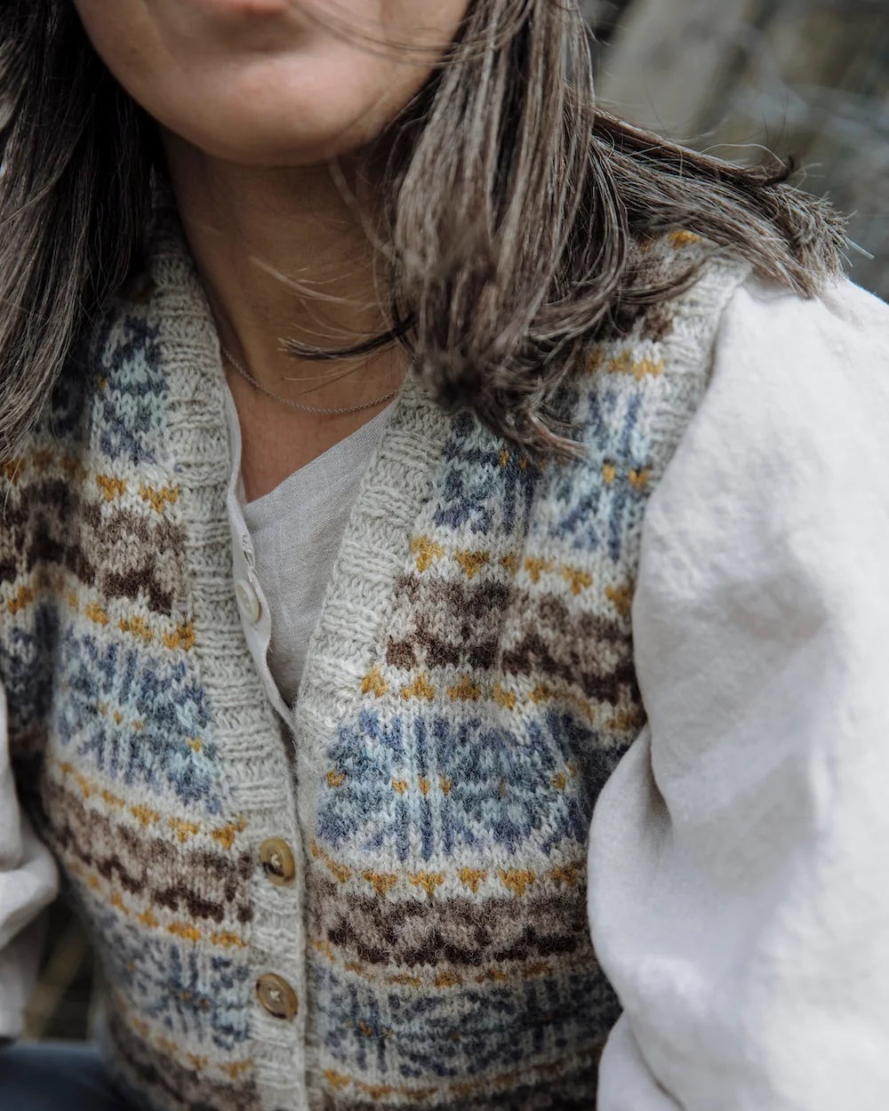A close-up of a person wearing a cream-colored knit button-front vest with blue, brown, and mustard geometric patterns, layered over a white shirt.