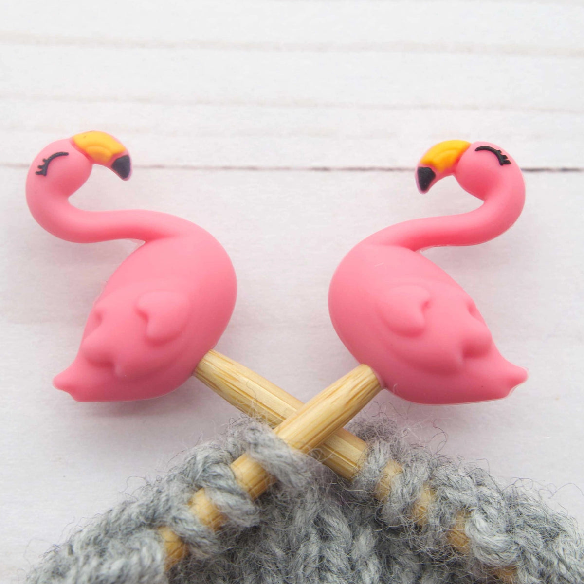 Two pink flamingo-shaped knitting needles with yellow and black beaks, shown inserted into gray yarn on a light wood background.