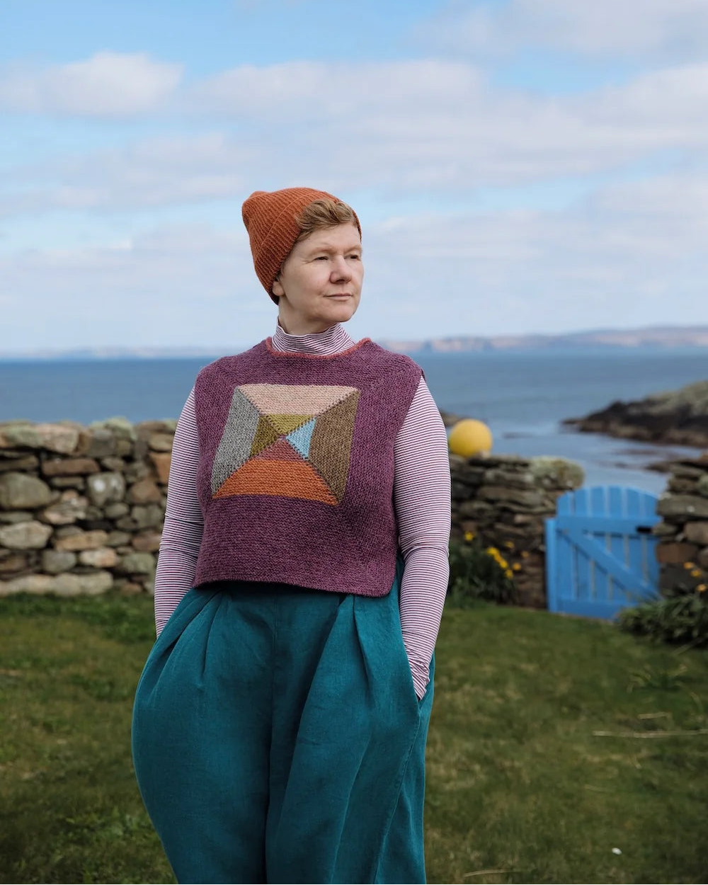 A woman standing outdoors by the sea wearing a textured geometric patterned sweater vest over a light pink long-sleeve top and teal wide-leg trousers, with an orange knit beanie. The background features a stone wall, grass, a blue gate, and a cloudy blue sky.