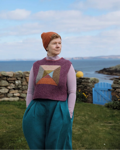 A woman standing outdoors by the sea wearing a textured geometric patterned sweater vest over a light pink long-sleeve top and teal wide-leg trousers, with an orange knit beanie. The background features a stone wall, grass, a blue gate, and a cloudy blue sky.
