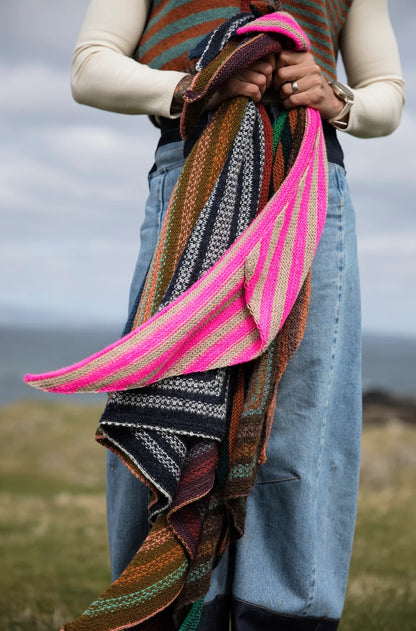 A person outdoors holding a large, vibrant handwoven shawl with multiple colors and patterns, including bright pink, green, brown, black, and white.