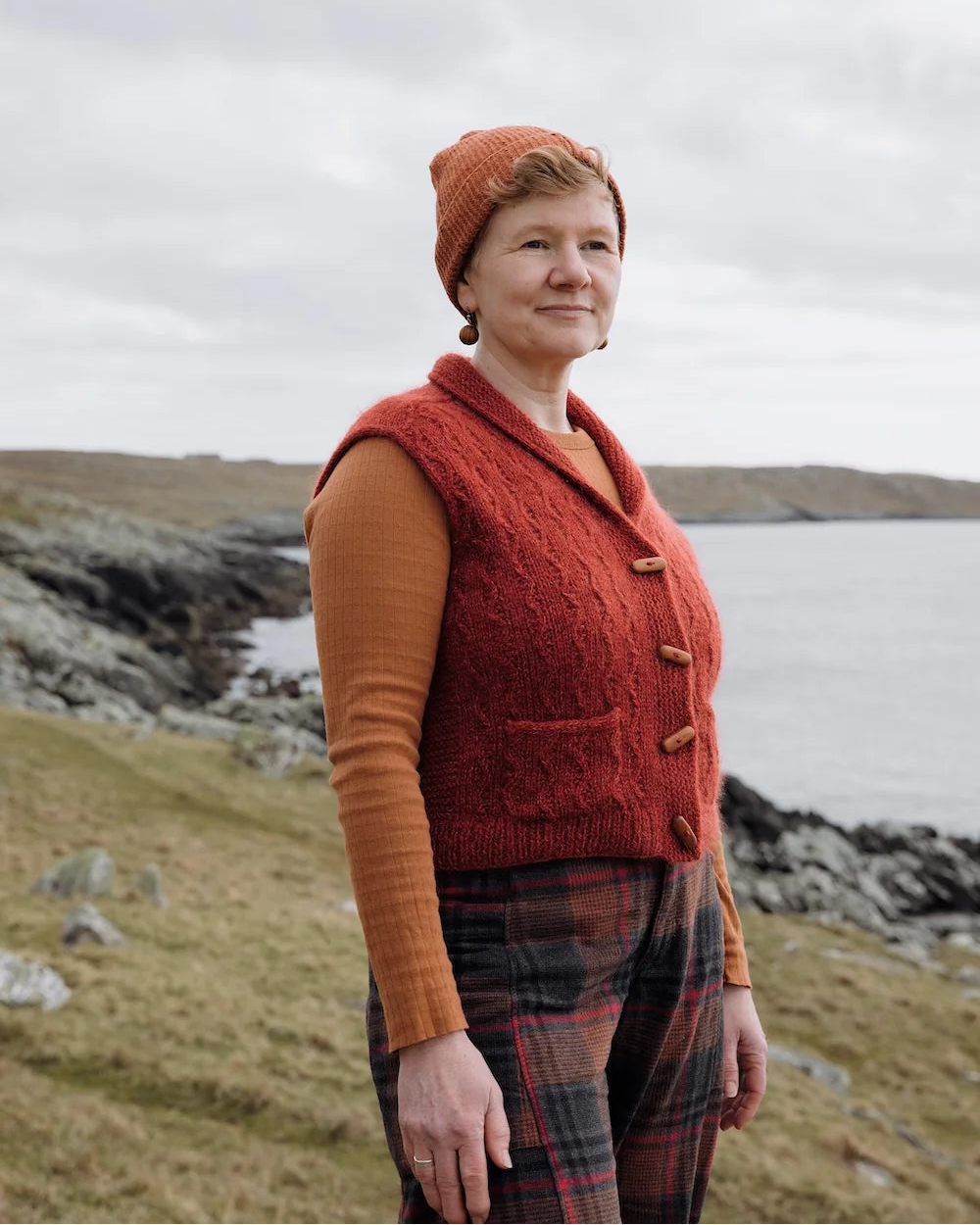 A woman standing outdoors on a grassy coastal hill under an overcast sky, wearing a rust-orange cable-knit vest over a rust-orange long-sleeve top, with plaid trousers.