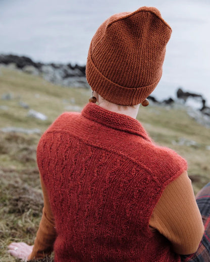 A person sitting outdoors on grassy ground, shown from behind, wearing a rust-orange knit beanie, a matching textured knit sweater vest over a rust-orange long-sleeve top, and plaid pants. The background shows a blurred natural landscape with water and rocks.