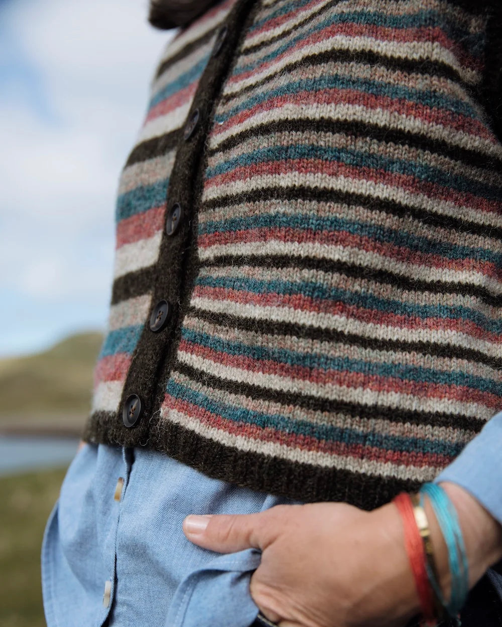 A close-up of a person wearing a striped knit cardigan with dark buttons over a light blue button-up shirt and dark jeans, with a blurred outdoor background.