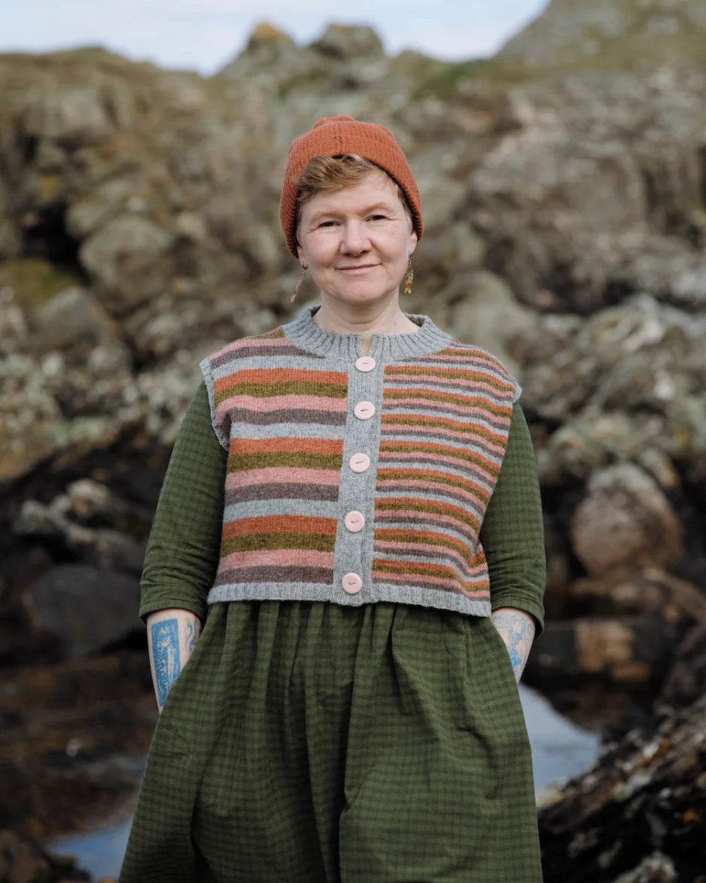 A woman standing outdoors on a rocky shoreline wearing a handmade knit outfit consisting of a striped cardigan vest over a green dress and an orange-brown beanie.