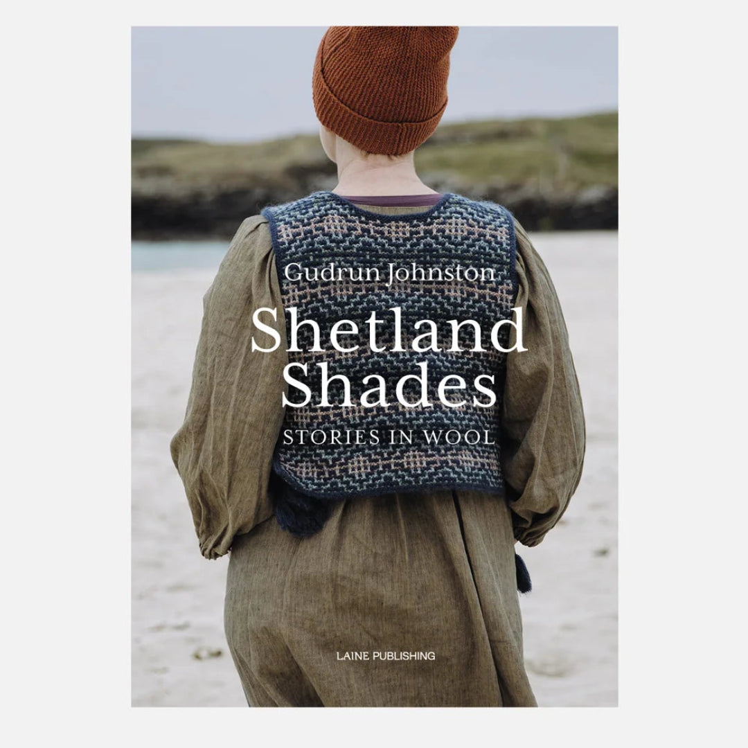 Book cover for 'Shetland Shades: Stories in Wool' by Gudrun Johnston, published by Lane Publishing. The cover features a photograph of a person from behind wearing a brown knit beanie and a patterned blue-and-white wool sweater vest over an olive green dress, standing on a beach.