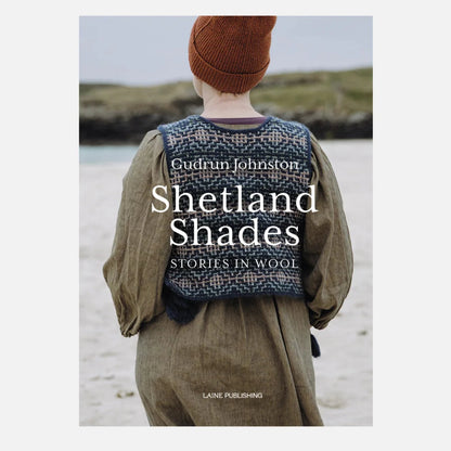 Book cover for 'Shetland Shades: Stories in Wool' by Gudrun Johnston, published by Lane Publishing. The cover features a photograph of a person from behind wearing a brown knit beanie and a patterned blue-and-white wool sweater vest over an olive green dress, standing on a beach.