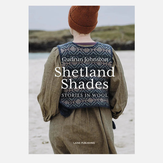 Book cover for 'Shetland Shades: Stories in Wool' by Gudrun Johnston, published by Lane Publishing. The cover features a photograph of a person from behind wearing a brown knit beanie and a patterned blue-and-white wool sweater vest over an olive green dress, standing on a beach.