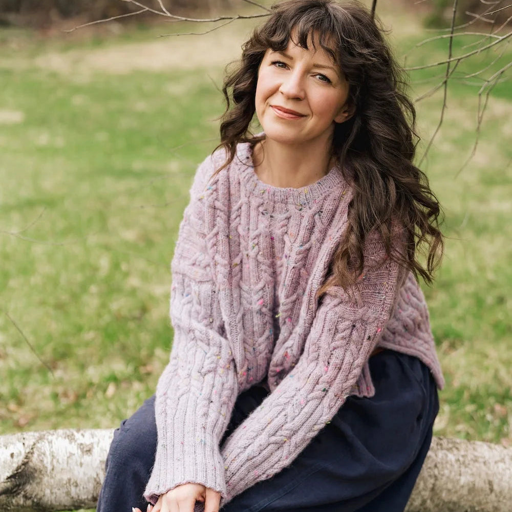 Woman sitting on a log in a natural setting wearing a pink sweater.