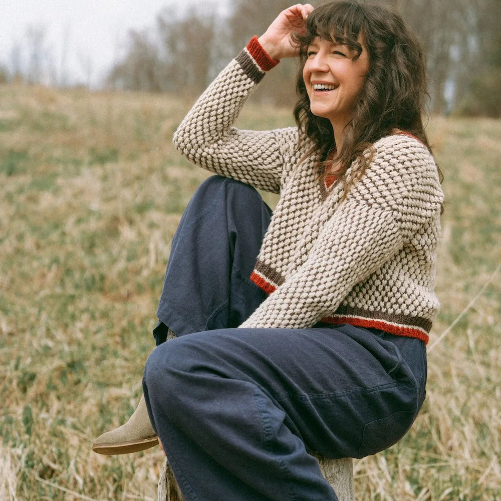 Woman sitting on a log in a field wearing a patterned sweater and blue pants.