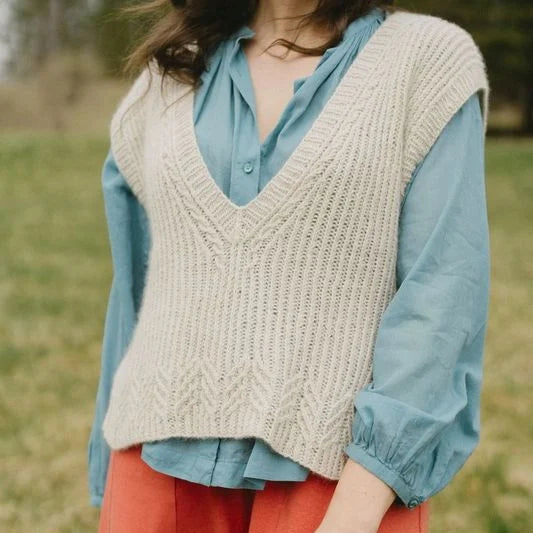 Person wearing a beige knitted vest over a blue shirt with red pants in a grassy outdoor setting