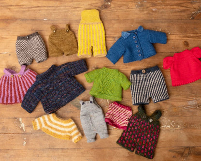 A collection of hand-knit doll clothes in various colors and patterns, including sweaters, shorts, and cardigans, arranged on a rustic wooden surface.
