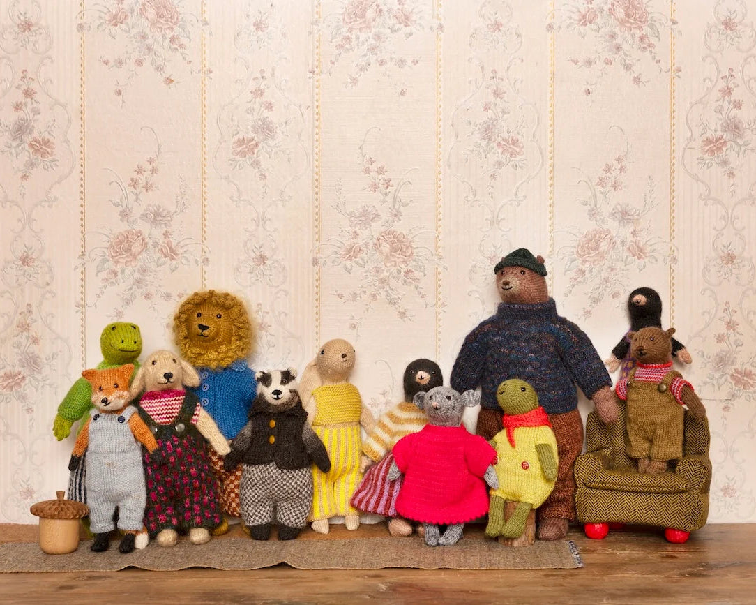 A group of handmade plush teddy bear figures and animal characters posed together on a wooden surface against a vintage floral wallpaper background.