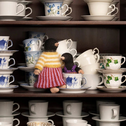 A collection of stacked white ceramic teacups and saucers on dark wooden shelves, with two small plush toy birds placed among the cups.